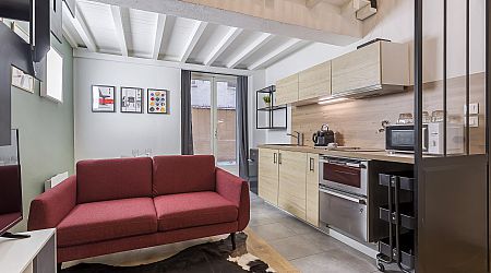 Studio moderne avec un canapé rouge, une petite cuisine avec des armoires en bois clair, des appareils électroménagers en acier inoxydable, des œuvres d'art murales et de grandes fenêtres laissant entrer la lumière naturelle. | 2BeApart Lyon