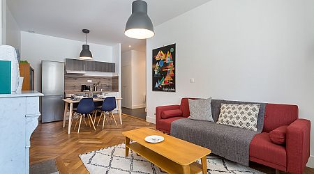 Salon moderne avec un canapé rouge, des coussins à motifs, une table basse en bois, un tapis, une table à manger avec quatre chaises bleues, et un coin cuisine avec des armoires grises et des appareils électroménagers en acier inoxydable en arrière-plan. | 2BeApart Lyon