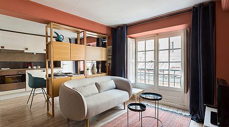 Un salon moderne avec un canapé gris clair, des tables basses rondes noires, un tapis rouge, une cloison en bois et de grandes fenêtres avec des rideaux sombres. La cuisine et la salle à manger sont visibles à l'arrière-plan. | 2BeApart Lyon