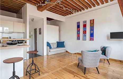 Salon moderne avec parquet et poutres apparentes au plafond, composé d'un petit canapé, de deux fauteuils à motifs, d'une télévision murale, d'œuvres d'art murales colorées et d'un bar de cuisine avec deux tabourets. | 2BeApart Lyon