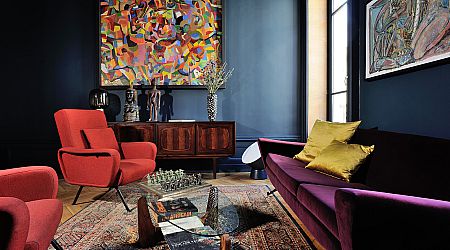 Un salon élégant avec des murs bleu foncé, des peintures abstraites, un fauteuil rouge, un canapé violet avec des coussins jaunes, un buffet en bois et une table basse en verre sur un tapis à motifs. | 2BeApart Lyon