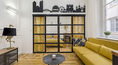 Un salon moderne avec un canapé jaune, une cloison en verre avec des cadres noirs et un décalque mural de la ligne d'horizon de la ville. Le sol en bois, le tapis rond et les grandes fenêtres laissent entrer la lumière naturelle. Une chambre à coucher est visible à travers la cloison. | 2BeApart Lyon
