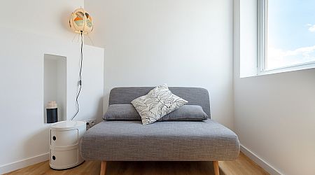 Pièce minimaliste avec un petit canapé gris, deux coussins à motifs, une table d'appoint ronde et blanche, une lampe murale moderne, un plancher en bois et une grande fenêtre laissant entrer la lumière naturelle. | 2BeApart Lyon