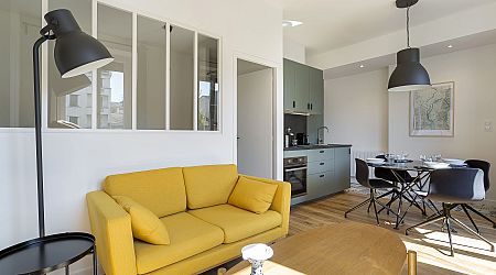 Salon moderne et lumineux avec un canapé jaune, un lampadaire noir et une petite table ronde, ouvrant sur une cuisine avec des armoires vertes et un coin repas avec une table ronde et des chaises noires. | 2BeApart Lyon