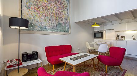 Salon moderne avec canapé et chaises rouge vif, table basse blanche, lampadaire et art abstrait au mur. Cuisine ouverte avec armoires blanches et table à manger visible en arrière-plan. | 2BeApart Lyon