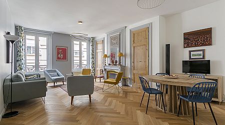 Salon et salle à manger modernes avec sols en bois à chevrons, canapés gris, chaises bleues autour d'une table en bois, fauteuil jaune, grandes fenêtres avec rideaux, télévision et œuvres d'art sur les murs. | 2BeApart Lyon