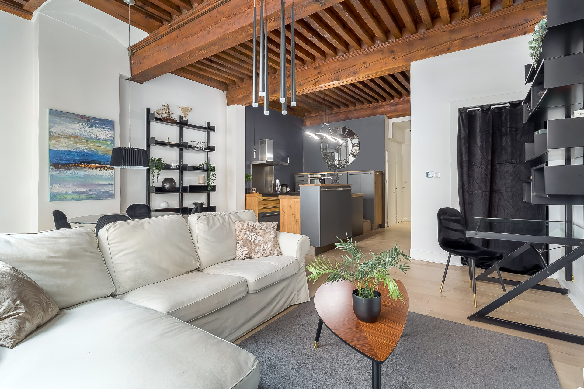Un salon moderne avec un canapé sectionnel beige, des poutres en bois au plafond, des étagères ouvertes et un mur d'accent noir - idéal pour les propriétaires exigeants qui recherchent une location meublée à Lyon. Une plante est posée sur la table basse près de la salle à manger. | 2BeApart Lyon