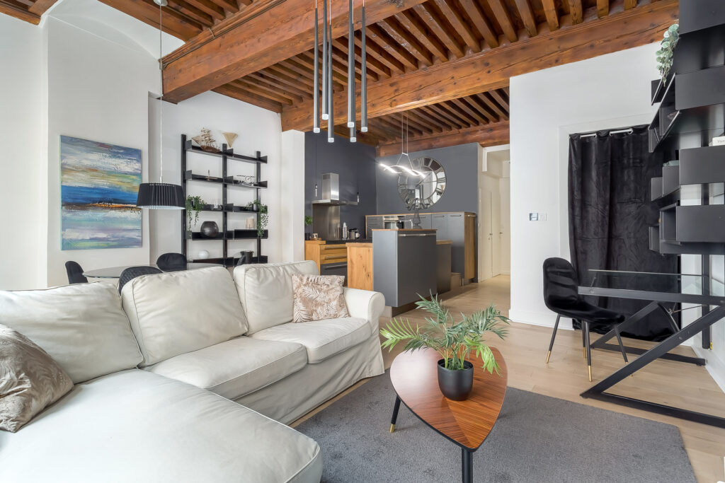 A modern living room in a location meublée Lyon with a white sectional sofa, wooden ceiling beams, open shelving with plants, a round mirror, small kitchen area, and a black desk with a chair against a white wall. | 2BeApart Lyon
