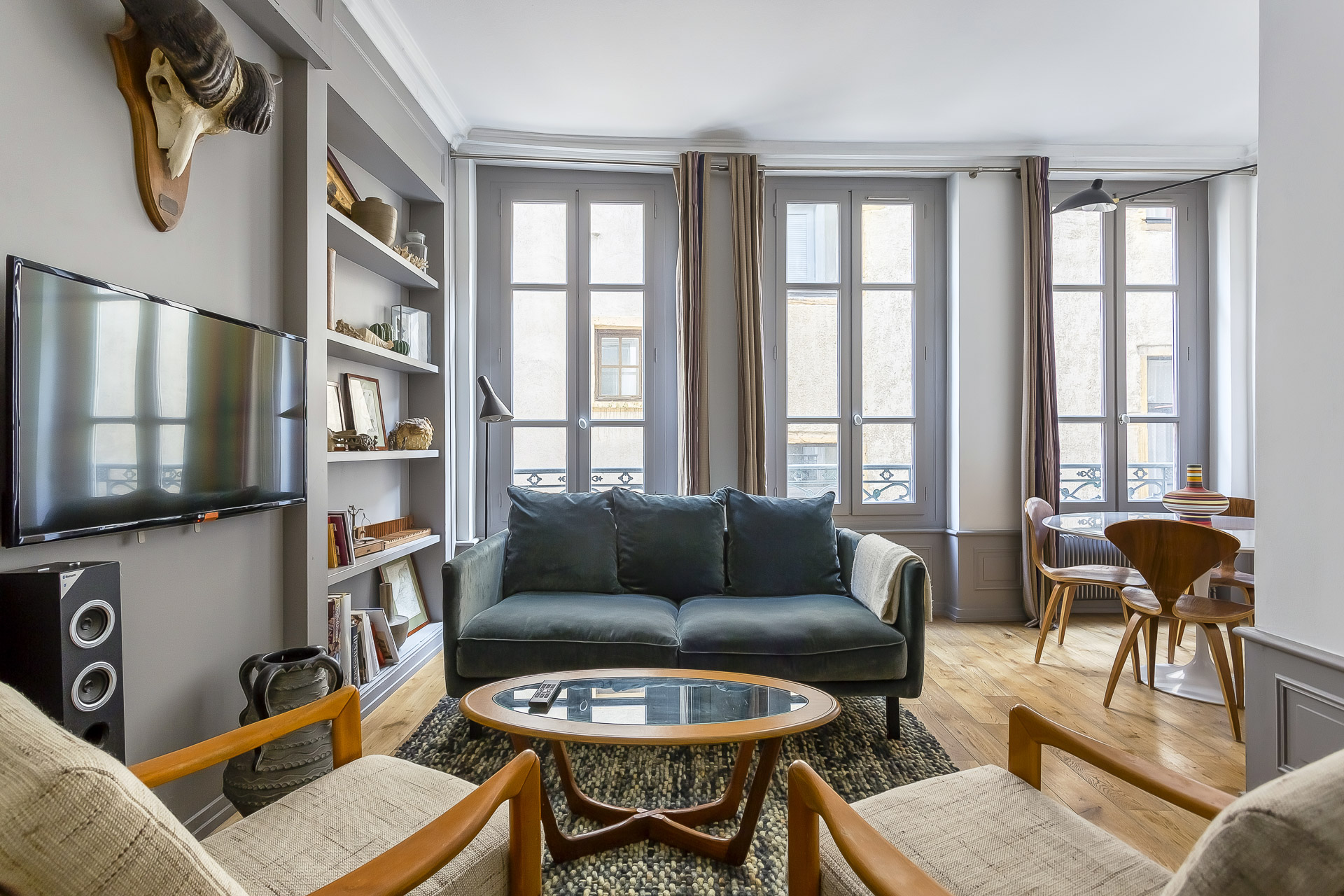 Bright living room with large windows, a dark green sofa, two armchairs, a round coffee table, wall-mounted TV, bookshelves, wooden floors, and a dining area—perfect for moyenne durée stays or a conciergerie haut de gamme experience. | 2BeApart Lyon