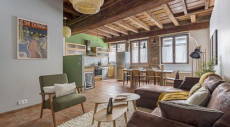 Salon et cuisine ouverts et confortables avec des poutres en bois rustiques, un canapé sectionnel marron, un fauteuil vert, une table de salle à manger, des appareils modernes et de grandes fenêtres laissant entrer la lumière naturelle. | 2BeApart Lyon