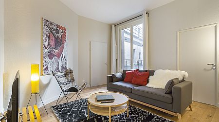 Salon moderne avec un canapé gris, des coussins rouges et blancs, une table basse ronde, un tapis à motifs, un fauteuil papillon, un lampadaire jaune, des œuvres d'art murales abstraites et une grande fenêtre laissant entrer la lumière naturelle. | 2BeApart Lyon