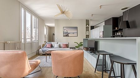 Salon moderne avec des planchers en bois clair, des fauteuils pêche, un canapé gris avec des coussins, une télévision sur un meuble bas, des tabourets de bar près d'un comptoir de cuisine, de grandes fenêtres et des œuvres d'art murales. L'espace est lumineux et aéré. | 2BeApart Lyon