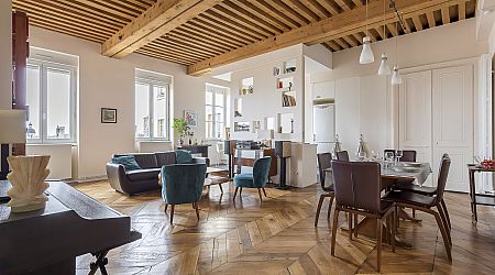 Un salon et une salle à manger spacieux et modernes avec des sols en bois, des poutres apparentes au plafond, des meubles contemporains, de grandes fenêtres et des murs aux tons neutres. La pièce est lumineuse et décorée avec style. | 2BeApart Lyon