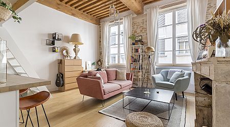 Salon lumineux avec des poutres en bois, de grandes fenêtres, un canapé rose, un fauteuil bleu, une bibliothèque et une décoration moderne. Le parquet en bois clair, les briques apparentes et les tons neutres confèrent à l'espace une atmosphère à la fois confortable et élégante. | 2BeApart Lyon
