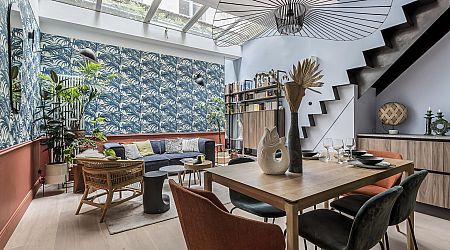 Salon et salle à manger modernes avec papier peint à motifs, plafond en verre, plantes, meubles éclectiques et étagères ouvertes. Une table en bois est dressée pour les repas près d'un escalier à la rampe en métal noir. | 2BeApart Lyon