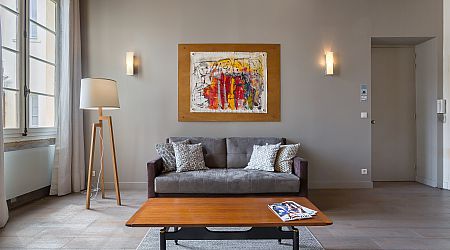 Salon moderne avec un canapé gris, des coussins à motifs, une table basse en bois avec des magazines, un lampadaire, de l'art mural abstrait et des murs et sols de couleur neutre. Une grande fenêtre laisse entrer la lumière naturelle. | 2BeApart Lyon