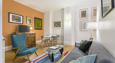 Un salon confortable avec un canapé gris, un fauteuil d'appoint bleu et un tapis rayé coloré. Un mur d'accent orange accueille des œuvres d'art encadrées, une petite télévision est posée sur un meuble en bois et une table de salle à manger avec quatre chaises se trouve à proximité. | 2BeApart Lyon