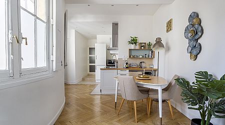 Cuisine et salle à manger d'un appartement moderne et lumineux avec des sols en bois, des murs blancs, une petite table avec deux chaises, des plantes vertes et une grande fenêtre qui laisse entrer la lumière naturelle. | 2BeApart Lyon