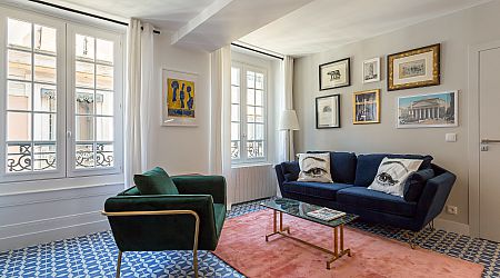 Salon lumineux avec de grandes fenêtres, un sol à motifs bleus, un canapé bleu avec des coussins imprimés, un fauteuil vert, un tapis rose, une table basse et un mur décoré d'œuvres d'art et de photos encadrées. | 2BeApart Lyon