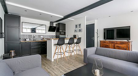 Espace de vie moderne ouvert avec un petit coin cuisine, des armoires noires, un bar de petit-déjeuner avec trois tabourets noirs, un canapé gris, un meuble TV en bois et un téléviseur mural. La décoration est minimaliste et élégante. | 2BeApart Lyon