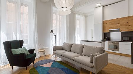 Salon moderne avec un canapé gris clair, un fauteuil noir, un tapis rond coloré, un lampadaire et une cuisine ouverte avec des armoires blanches et des accents en bois. De grandes fenêtres avec des rideaux diaphanes laissent entrer la lumière naturelle. | 2BeApart Lyon