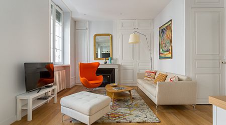 Salon lumineux avec un canapé beige, un fauteuil d'appoint orange, un ottoman blanc et une télévision sur pied. Un tapis moderne, un tableau mural abstrait et un grand miroir au-dessus de la cheminée complètent cet espace confortable et élégant. | 2BeApart Lyon