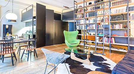 Espace de vie moderne avec un fauteuil d'appoint vert, une table basse en verre sur un tapis en peau de vache, des étagères ouvertes remplies de livres et un coin repas avec des chaises noires. Le sol est en bois clair et la décoration est contemporaine. | 2BeApart Lyon