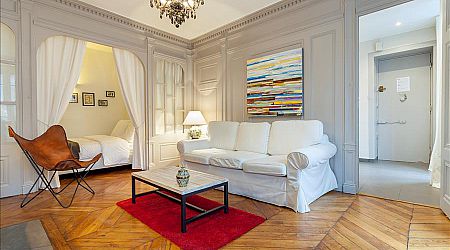 Salon élégant avec un canapé blanc, des œuvres d'art modernes, des planchers en bois et un tapis rouge. Une chambre à coucher en alcôve avec des rideaux et un lit blancs est visible sur la gauche, et un lustre élégant est suspendu au plafond. | 2BeApart Lyon