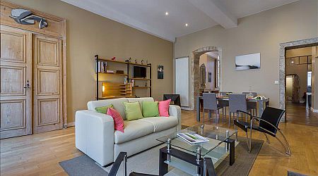 Salon moderne avec un canapé blanc, des coussins colorés, une table basse en verre, deux chaises noires, des étagères en bois et un coin repas en arrière-plan. Les planchers en bois clair et les murs neutres créent un espace confortable et accueillant. | 2BeApart Lyon