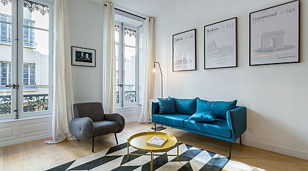 Un salon lumineux avec de grandes fenêtres, un canapé sarcelle, un fauteuil gris, une table basse ronde jaune, un tapis géométrique et des œuvres d'art encadrées au mur. La lumière du soleil remplit l'espace moderne avec des planchers en bois clair. | 2BeApart Lyon
