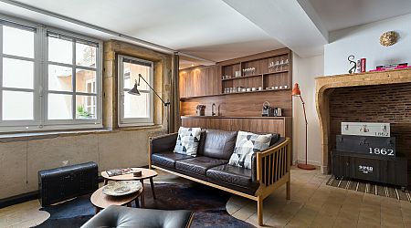 Un salon confortable avec un canapé moderne en bois, des coussins en cuir noir, des oreillers à motifs, une table basse ronde, une malle noire, des étagères encastrées avec des verres, et une grande cheminée rustique près d'une fenêtre. | 2BeApart Lyon
