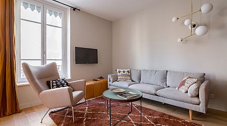 Salon moderne avec un canapé gris, des coussins à motifs, un fauteuil clair, une table basse ronde en verre, un tapis marron, une télévision murale et un lustre contemporain. Une grande fenêtre avec des rideaux dorés laisse entrer la lumière naturelle. | 2BeApart Lyon