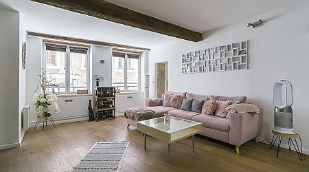 Un salon moderne avec des planchers en bois, un canapé sectionnel rose pâle avec des coussins, une table basse blanche, des œuvres d'art murales, de grandes fenêtres et un décor minimaliste comprenant des plantes et un ventilateur. | 2BeApart Lyon
