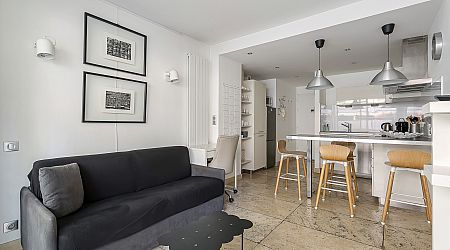 Espace de vie moderne et lumineux avec un canapé noir, deux tableaux encadrés au mur, un îlot de cuisine avec des tabourets en bois, des lampes suspendues et une cuisine blanche minimaliste en arrière-plan. | 2BeApart Lyon