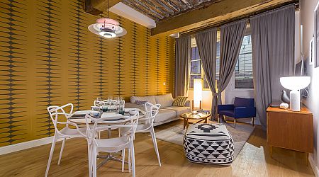 Salon moderne avec papier peint rétro à motifs jaunes, poutres en bois apparentes, rideaux gris, table à manger ronde avec chaises blanches, canapé beige, fauteuil bleu, ottoman géométrique et éclairage chaleureux. | 2BeApart Lyon