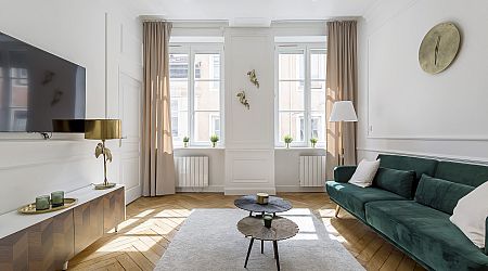 Salon lumineux avec de grandes fenêtres, des rideaux beiges, un canapé en velours vert, des tables basses rondes, une télévision au mur, une décoration moderne et des sols en bois clair. Ambiance minimaliste, élégante et aérée. | 2BeApart Lyon