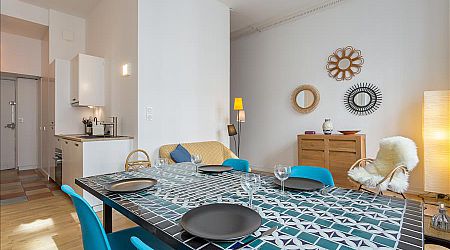 Une salle à manger et un salon modernes avec une table à motifs géométriques, des chaises bleues, un canapé clair et un buffet en bois. Des miroirs et des lampes décoratives ajoutent une touche de confort à cet espace ouvert et lumineux. | 2BeApart Lyon