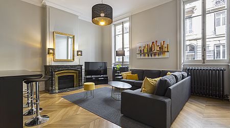 Salon moderne avec canapé sectionnel gris, accents jaunes, table basse ronde, cheminée avec miroir encadré d'or, télévision à écran plat, grandes fenêtres et sol en bois à chevrons. Des œuvres d'art contemporain sont accrochées au mur. | 2BeApart Lyon