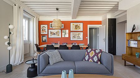 A modern living-dining room features a gray sofa with patterned cushions, a round wooden coffee table, a black floor lamp, and a dining table with black chairs. The back wall is orange with framed art and a pendant light. | 2BeApart Lyon