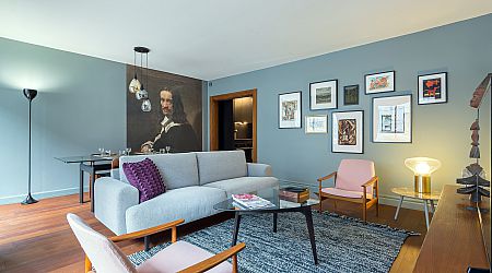 Salon moderne avec des murs bleu clair, un canapé gris, deux fauteuils roses, une table basse et des œuvres d'art murales. Un coin repas avec un portrait peint se trouve à l'arrière-plan ; un tapis et un lampadaire ajoutent de la chaleur à l'espace. | 2BeApart Lyon