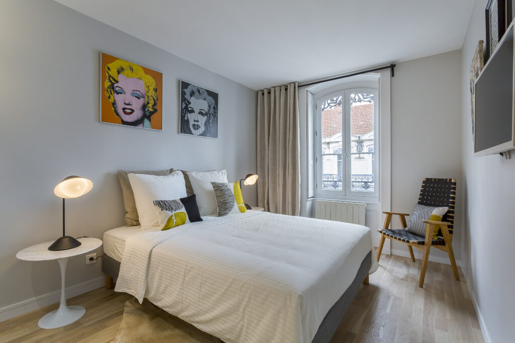 Une chambre moderne avec un lit double, des portraits pop art colorés au-dessus de la tête de lit, une table ronde avec une lampe, une chaise en bois et une grande fenêtre avec des rideaux gris - idéal pour les besoins de location meublée Lyon ou de gestion locative. | 2BeApart Lyon