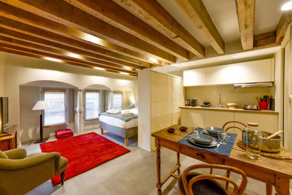 Studio douillet avec poutres apparentes, lit près de deux fenêtres, tapis rouge et petite cuisine. Cette charmante location meublée dispose d'une table à manger en bois pour deux personnes et d'un éclairage doux pour une atmosphère chaleureuse et accueillante. | 2BeApart Lyon