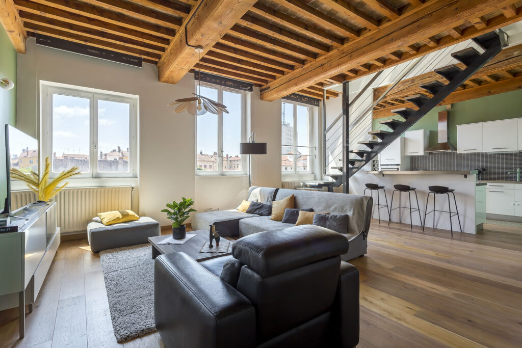 Appartement loft moderne location meublée avec des poutres en bois apparentes, de grandes fenêtres, un coin salon confortable avec des canapés gris, des plantes, une cuisine avec des armoires blanches, un bar de petit déjeuner avec des tabourets, et un escalier en métal noir. | 2BeApart Lyon