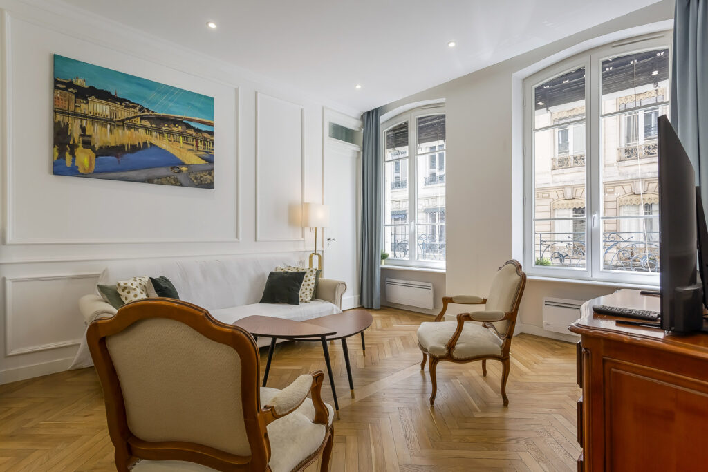 Un salon lumineux dans une location meublée à Lyon comprend un canapé blanc, deux fauteuils rembourrés, une table basse en bois, une télévision et de grandes fenêtres. Un tableau coloré est accroché au-dessus du canapé et la lumière du soleil entre à flots. Conciergerie haut de gamme incluse. | 2BeApart Lyon