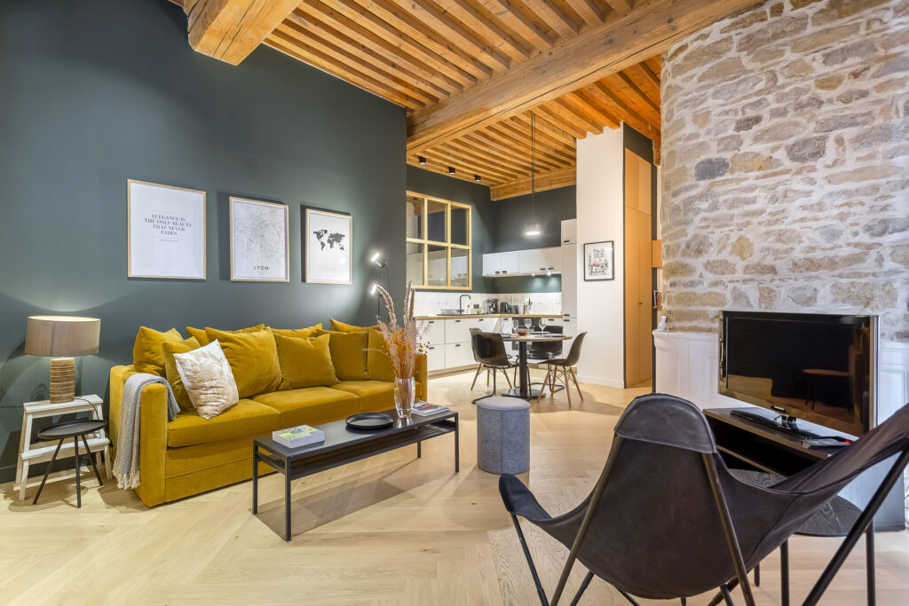 Un salon moderne et confortable avec un canapé jaune moutarde, une table basse noire, des œuvres d'art murales et un mur d'accent en pierre. Parfait pour les propriétaires exigeants, cet espace présente des plafonds en poutres apparentes, un parquet en bois clair et une cuisine ouverte avec un coin repas. | 2BeApart Lyon