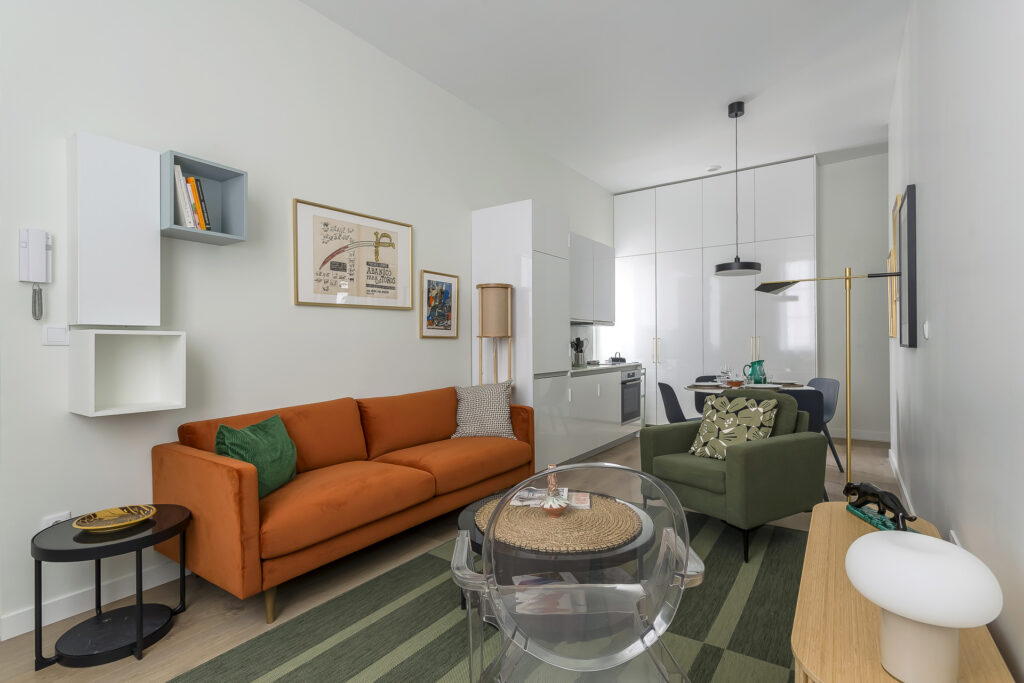 Salon moderne avec un canapé orange, un fauteuil vert, un petit coin repas et une cuisine blanche. Parfait pour les propriétaires exigeants, l'espace comprend des œuvres d'art murales, des étagères, une table basse en verre posée sur un tapis à rayures vertes et une lumière naturelle intense. | 2BeApart Lyon