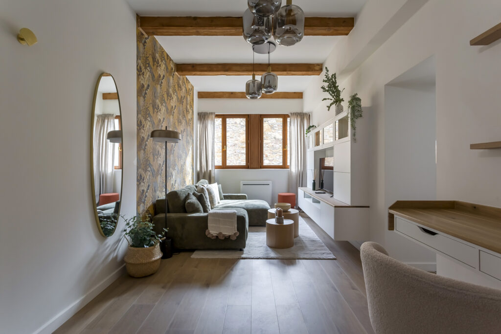 Un salon moderne avec des poutres en bois, un canapé vert, un miroir rond, un mur d'accent à motifs, des étagères murales, une télévision et de grandes fenêtres laissant entrer la lumière naturelle. Parfait pour la location meublée Lyon, avec des tons neutres et des plantes qui ajoutent une touche de confort. | 2BeApart Lyon