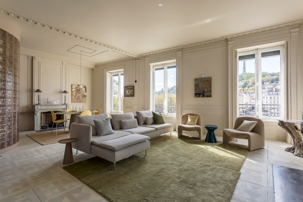 Un salon spacieux et élégant avec de grandes fenêtres, des murs clairs, des meubles modernes et un tapis vert offre une vue sur les collines et le paysage urbain, ce qui est parfait pour les propriétaires exigeants à la recherche d'une location meublée à Lyon. Un canapé, des chaises et une salle à manger complètent l'espace. | 2BeApart Lyon
