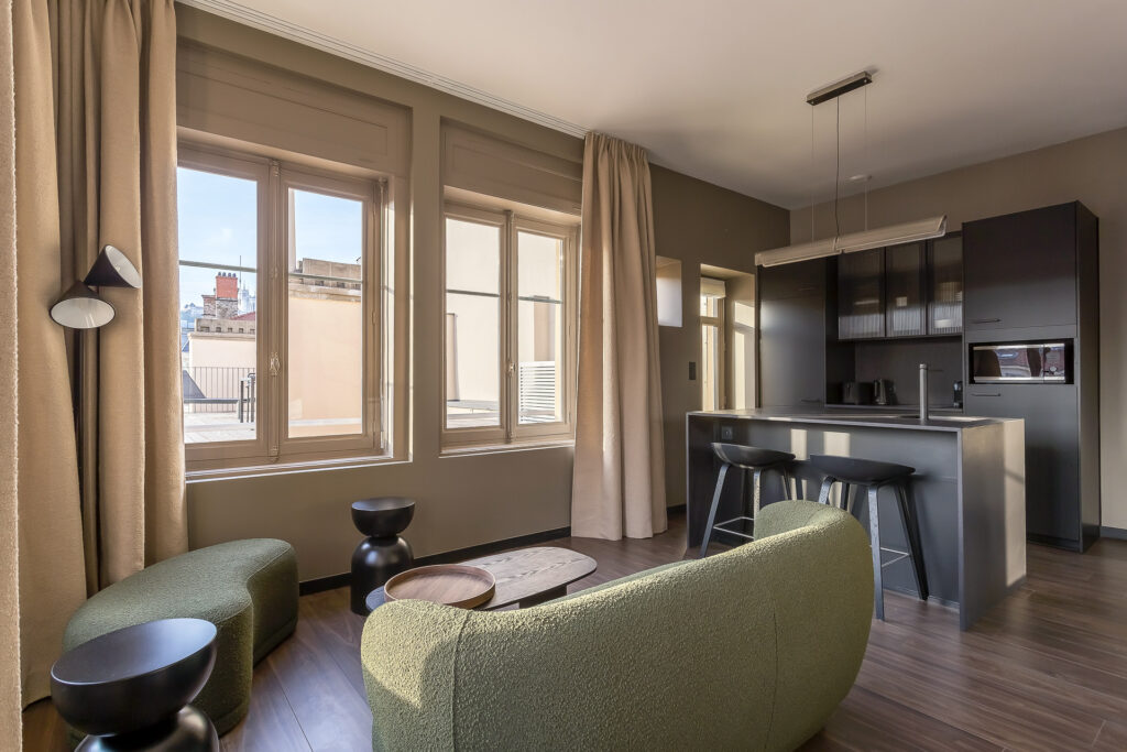Salon d'appartement moderne avec de grandes fenêtres, des canapés verts, des tables basses rondes, un sol en bois foncé et une cuisine ouverte avec des armoires noires et des tabourets de bar. Idéal pour les propriétaires exigeants à la recherche de confort et de conciergerie haut de gamme. | 2BeApart Lyon