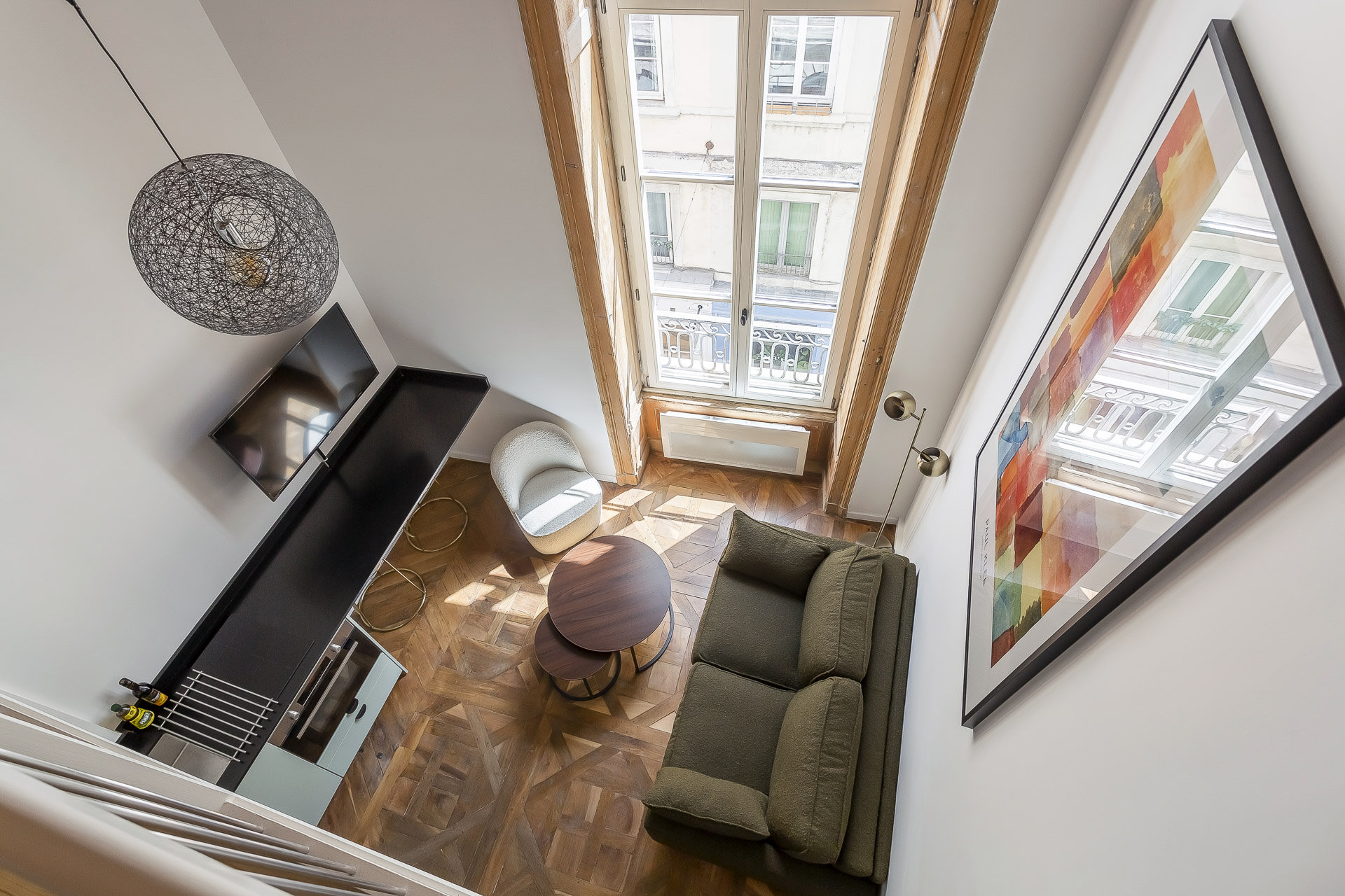 Un salon moderne vu d'en haut dans une location meublée à Lyon, avec un canapé vert, une table basse ronde, une télévision sur une console noire, de grandes fenêtres avec vue sur la ville, un parquet en bois et une peinture abstraite colorée sur le mur. | 2BeApart Lyon